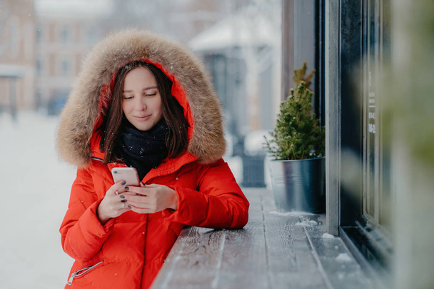 Kuva nuoresta naisesta punaisessa lämpimässä takissa, pitää nykyaikaista matkapuhelinta, lähettää tekstiviestin poikaystävälle, jakaa esteitä talvilomista, seisoo ulkona lumen aikana. Teknologian käsite
. - Valokuva, kuva