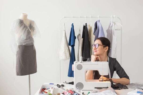 Kleermaker, naaister en mode concept - lachende vrouwelijke modeontwerper met behulp van de naaimachine en zittend achter haar Bureau - Foto, afbeelding