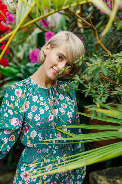 portrait of a young beautiful blonde woman in a green dress in a beautiful botanic garden with flowers - Fotoğraf, Görsel