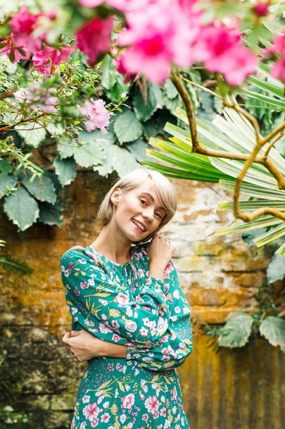 portrait of a young beautiful blonde woman in a green dress in a beautiful botanic garden with flowers - Fotó, kép