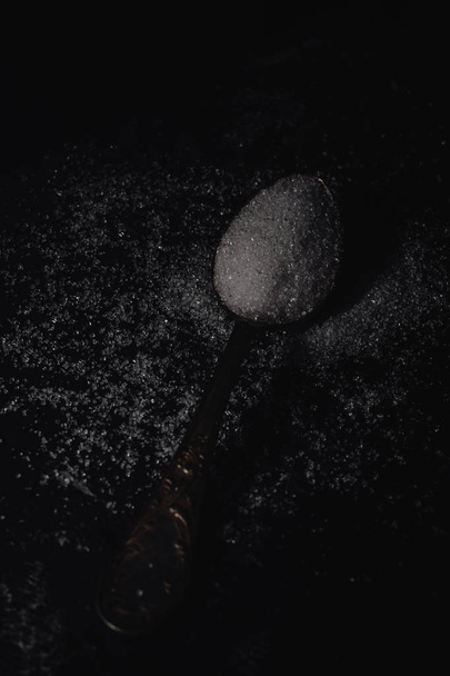 Tmavé fotografie lžíce bílého cukru na černém pozadí kamene s kopií prostor, vodorovný řez, nízké klíč osvětlení - Fotografie, Obrázek