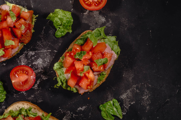 Traditional Italian Bruschetta with chopped tomatoes, mozzarella sauce, salad leaves and ham on a dark baton background. Traditional italian appetizer or snack, antipasto. Top view with copy space, co - Foto, afbeelding