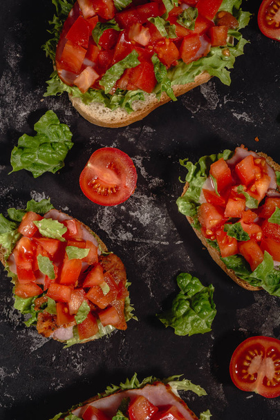 Traditional Italian Bruschetta with chopped tomatoes, mozzarella sauce, salad leaves and ham on a dark baton background. Traditional italian appetizer or snack, antipasto. - Photo, Image