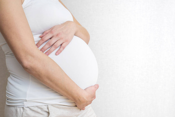 Terhes nő tartja kezében a hasa, a világos háttér. Terhesség, anyaság, előkészítése és elvárás koncepció. Közelkép, másol hely, zárt térben. Gyönyörű pályázati hangulat fénykép a terhesség - Fotó, kép