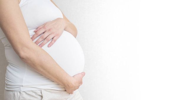 Terhes nő tartja kezében a hasa, a világos háttér. Terhesség, anyaság, előkészítése és elvárás koncepció. Közelkép, másol hely, zárt térben. Gyönyörű pályázati hangulat fénykép a terhesség - Fotó, kép