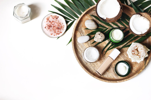 Styled beauty composition. Skin creams, makeup bottle, rose and pebble stones on wooden tray. Coconuts, tropical palm leaves decoration. Cosmetics, spa concept. Empty space, flat lay, top view. - Foto, imagen