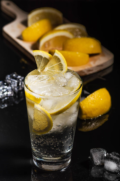 Gin tonic met plakjes citroen en ijsblokje, met citroen en ijs voor versieren op rug, op de zwarte achtergrond - Foto, afbeelding