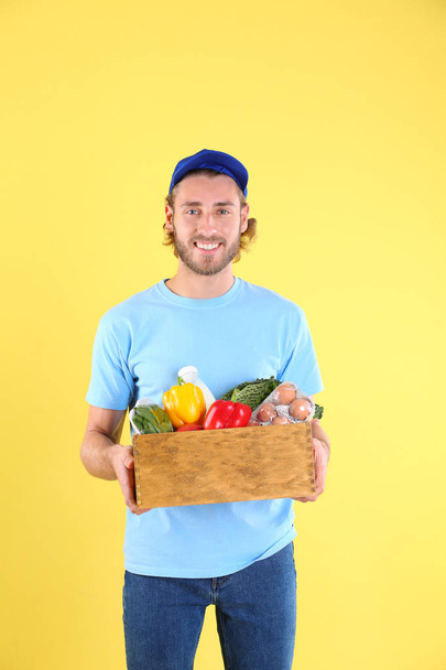 Delivery man holding wooden crate with food products on color background - Foto, immagini