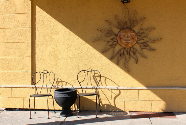 Mur en stuc peint en jaune avec une vieille décoration solaire en métal rustique suspendue sur des chaises et une table en métal noir au restaurant extérieur
 - Photo, image