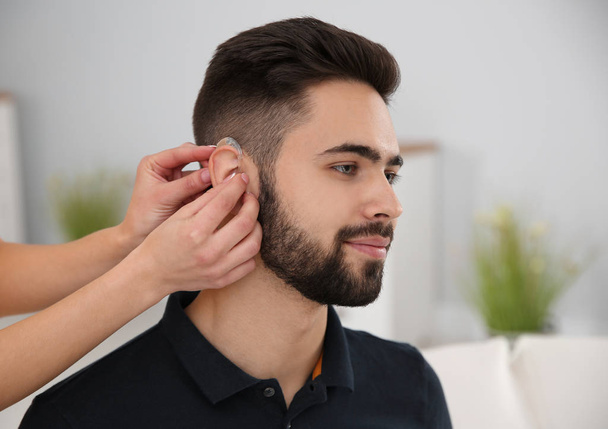 Young woman putting hearing aid in man's ear indoors - Foto, imagen