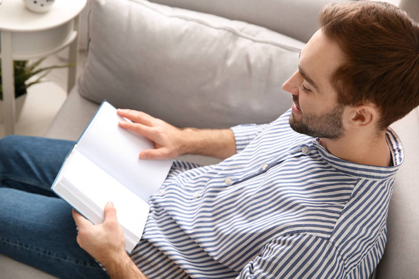 Handsome young man reading book on sofa at home - Foto, Bild