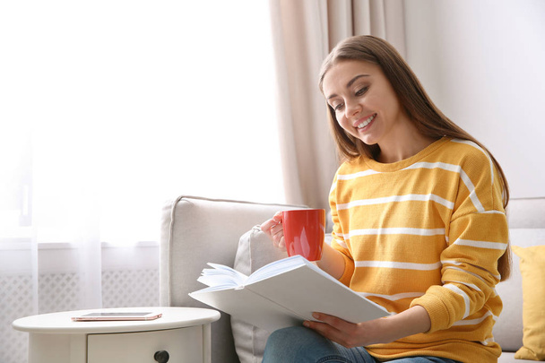 Young woman with cup of coffee reading book on couch at home - Photo, image