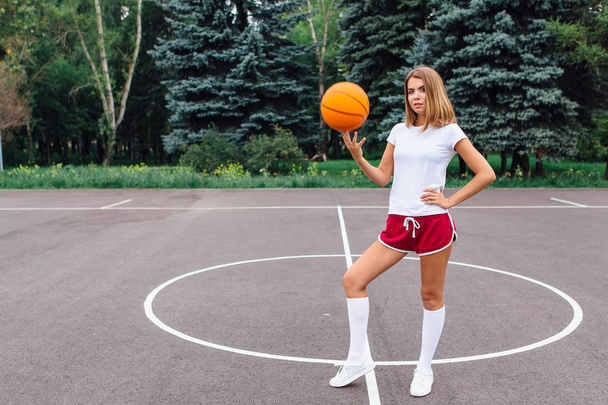 Beautiful young girl dressed in white t-shirt, shorts and sneakers, plays with a ball on a basketball court. - Фото, зображення