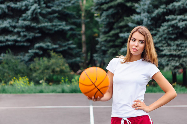 Beautiful young girl dressed in white t-shirt, shorts and sneakers, plays with a ball on a basketball court. - Fotoğraf, Görsel