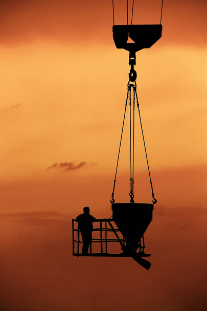 silhouette of a worker at a construction site, which rises on a crane together with a concrete mixer on the background of an orange sunset - Foto, Imagem