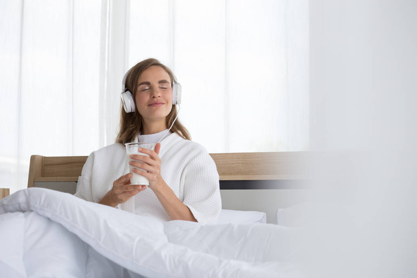 Frau hält Glas Milch in der Hand und lauscht morgens auf dem Bett im Zimmer der Eigentumswohnung Entspannungsliedern bei der Musikanwendung - Foto, Bild