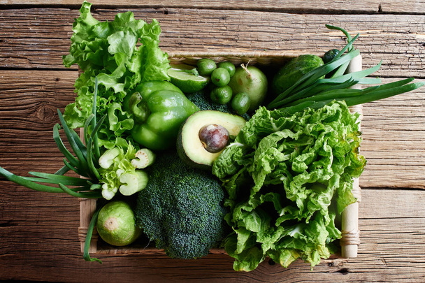 Green vegetables and fruits and greens in a brown wicker basket on a wooden background. Healthy eating concept - 写真・画像