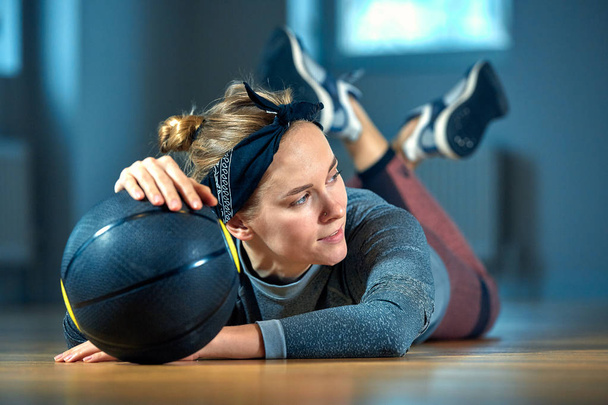 beautiful fit woman in sportswear posing while lying on the floor with basketball in front of window at gym Healthy girl lifestyle and sport concept - Photo, Image