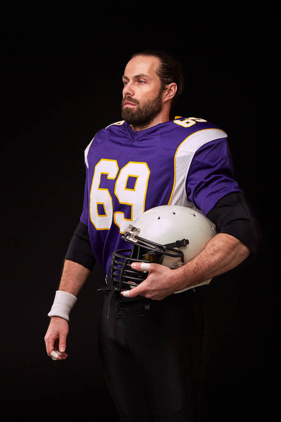 Configured for the game. American football player looking at camera and holding helmet in his hand while standing against black background - Fotoğraf, Görsel
