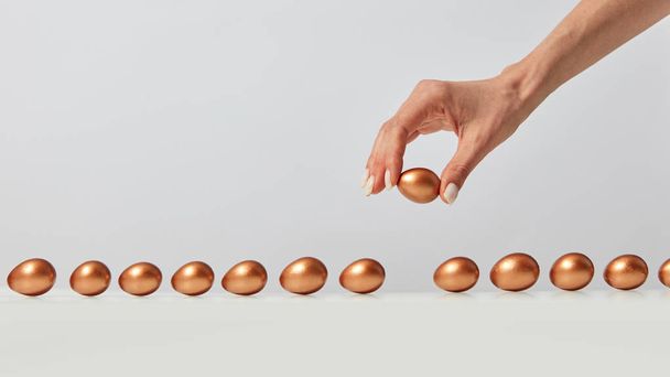 Woman's hand laying out a row of golden painted eggs on a gray background with copy space. The concept of wealth and profits - Фото, изображение