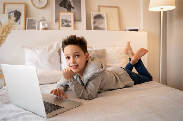 A little boy sitting on a big bed with a laptop book and looks into the camera. - Valokuva, kuva