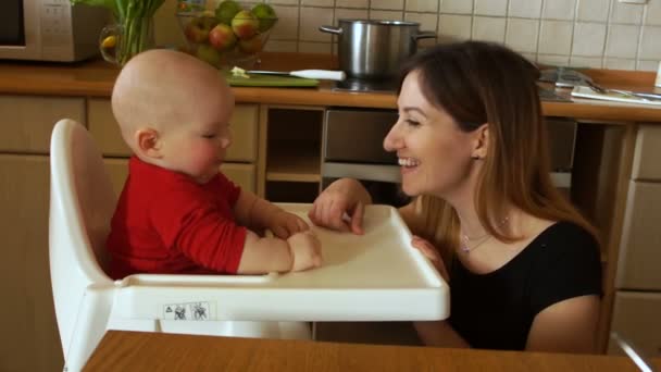 Young mother with baby in the kitchen. A woman feeds a baby sitting in a highchair. Lettuce leaf - Video