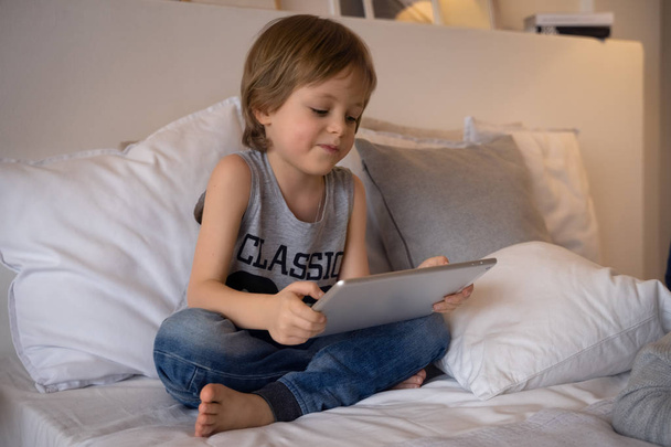 Menino de 5 anos deitado Na cama jogando jogo no tablet no quarto, Educação escola tecnologia internet conceito
 - Foto, Imagem