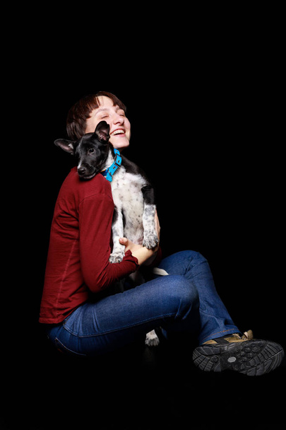 woman in red sweater and jeans sitting, hugging and holding mongrel dog in blue collar isolated on black - Φωτογραφία, εικόνα