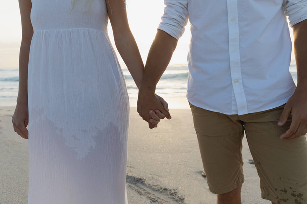 Mid sectie van jonge liefde paar bezit hand terwijl je op het strand op een zonnige dag. Ze genieten van hun vakantie - Foto, afbeelding
