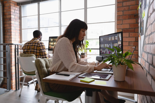 Zijaanzicht van de Kaukasische zakenvrouw werkt via laptop terwijl Afro-Amerikaanse man achter haar werken in office - Foto, afbeelding