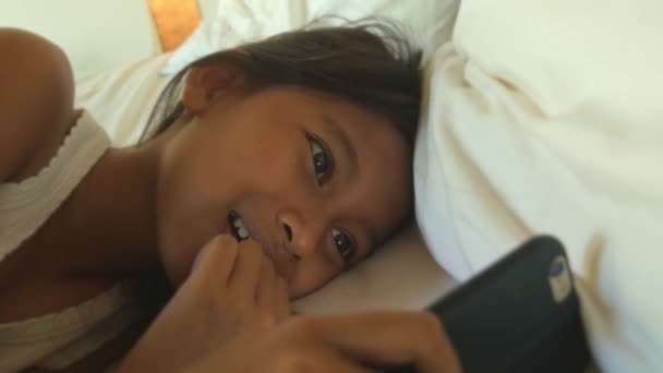 lifestyle close up head shot of sweet happy and beautiful 7 years old child having fun using internet mobile phone watching online lying on bed cheerful at night smiling cheerful - Footage, Video