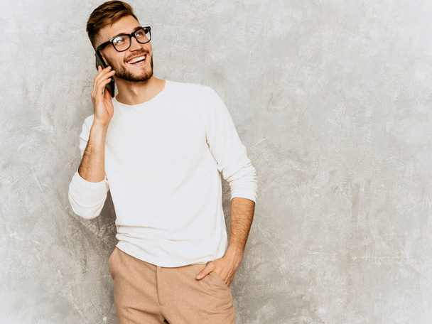 Portrait of handsome smiling hipster lumbersexual businessman model wearing casual summer white clothes. Fashion stylish man posing against gray wall. Speaking on mobile phone - 写真・画像