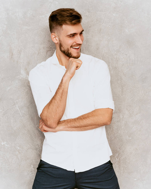 Портрет красивого улыбающегося хипстера-лесоруба модели бизнесмена, одетого в повседневную летнюю белую рубашку. Модный стильный мужчина позирует против серой стены
 - Фото, изображение