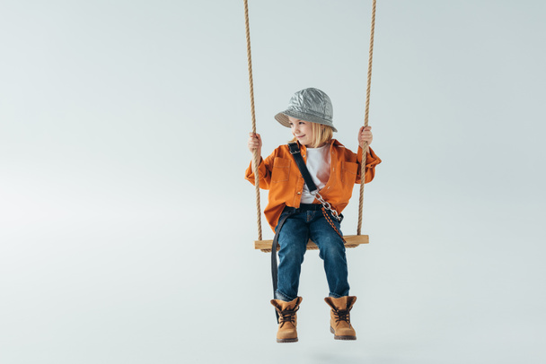 cute kid in jeans and orange shirt sitting on swing and looking away on grey background - Valokuva, kuva