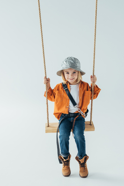 smiling kid in jeans and orange shirt sitting on swing on grey background - 写真・画像