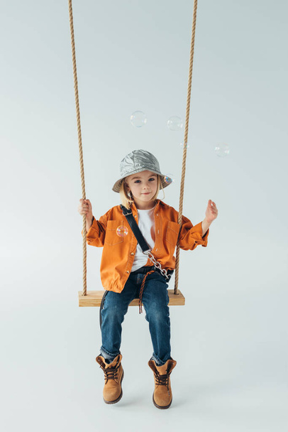 cute kid in jeans and orange shirt sitting on swing and looking at soap bubbles - Photo, image