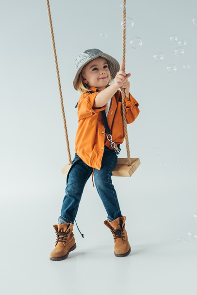 smiling kid in jeans and orange shirt sitting on swing and ponting with finger at soap bubbles - Photo, Image