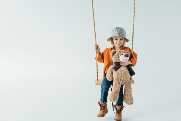 cute kid in jeans and orange shirt sitting on swing and holding teddy bear  - Fotó, kép