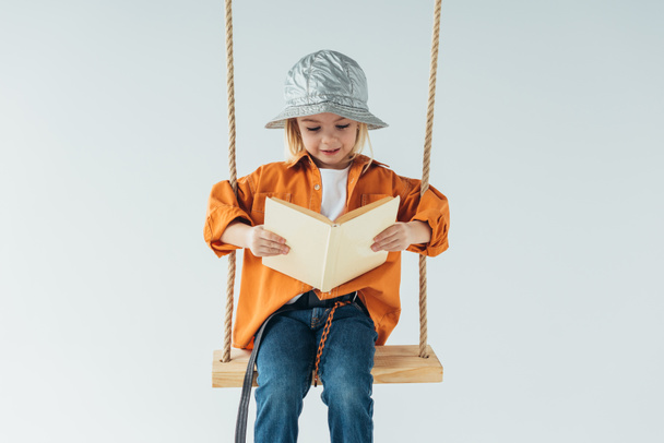 adorable kid in jeans and orange shirt sitting on swing and reading book isolated on grey - Photo, Image