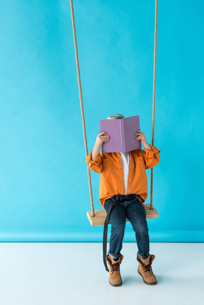 cute kid in jeans and orange shirt sitting on swing and reading book on blue background  - Фото, изображение