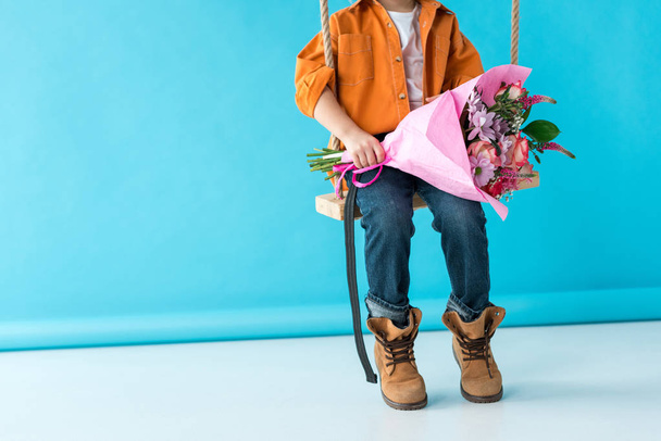 cropped view of kid sitting on swing and holding bouquet on blue background  - Φωτογραφία, εικόνα