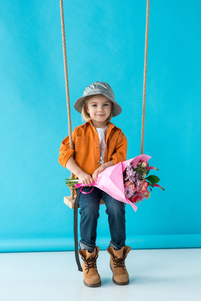 adorable kid sitting on swing, holding bouquet and looking at camera on blue background  - Photo, Image