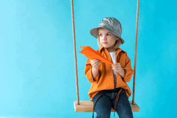 cute child sitting on swing, holding paper plane and looking away isolated on blue - Fotografie, Obrázek