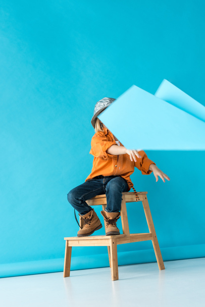 kid in jeans and orange shirt sitting on stairs and playing with paper plane  - Foto, Imagen