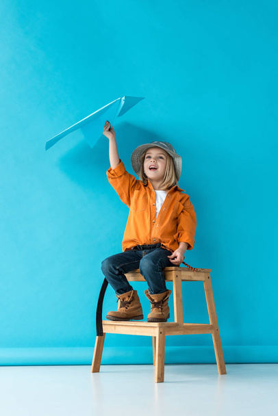 kid in jeans and orange shirt sitting on stairs and playing with blue paper plane  - Photo, image
