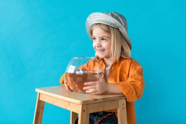 smiling kid in silver hat and orange shirt holding aquarium with goldfish isolated on blue - Foto, immagini
