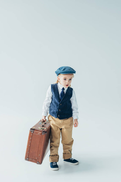 surprised kid in retro vest and cap holding suitcase on grey background  - Fotó, kép