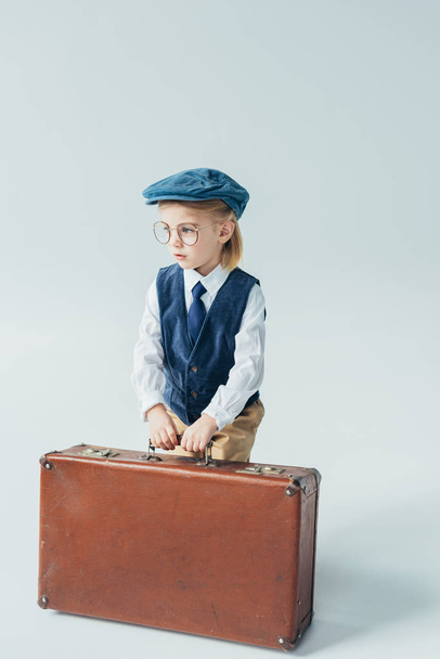 thoughtful kid in retro vest and cap holding suitcase on grey background  - Foto, afbeelding