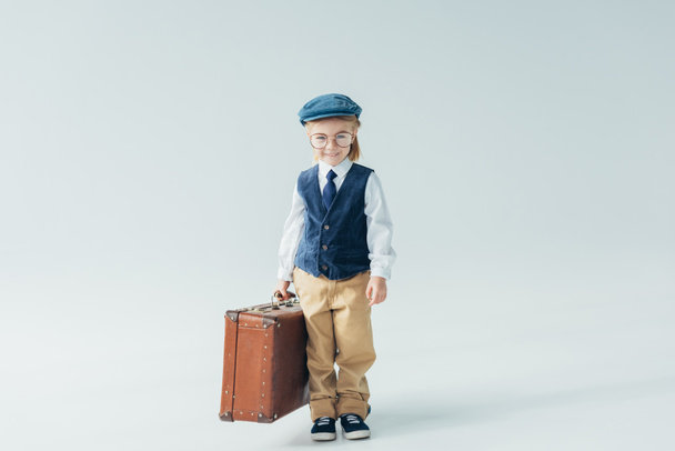Lächelndes Kind in Retro-Weste und Mütze mit Koffer auf grauem Hintergrund  - Foto, Bild
