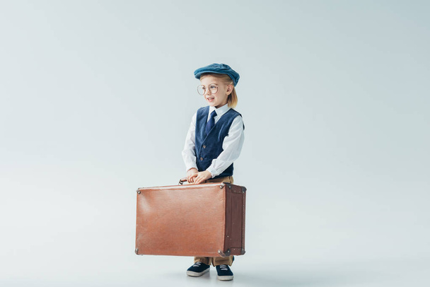 smiling kid in retro vest and cap holding suitcase and looking away on grey background  - Photo, Image